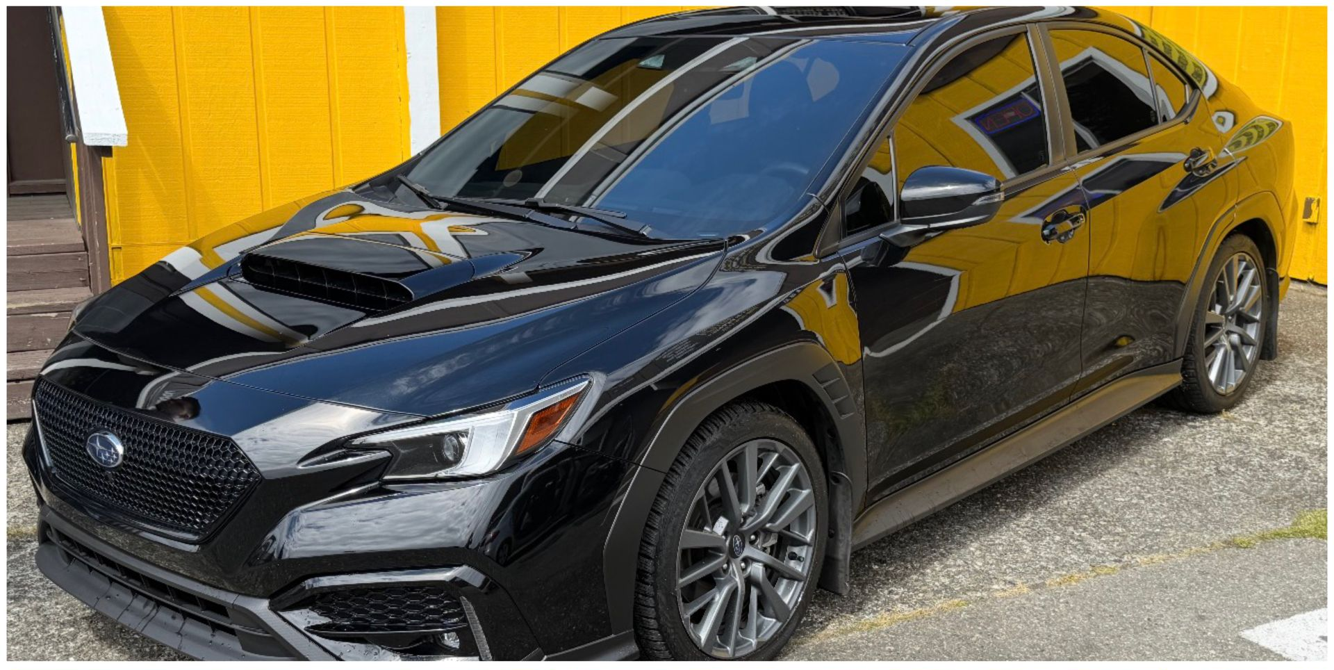 Black Subaru WRX parked in front of a yellow building with reflections in its windows.