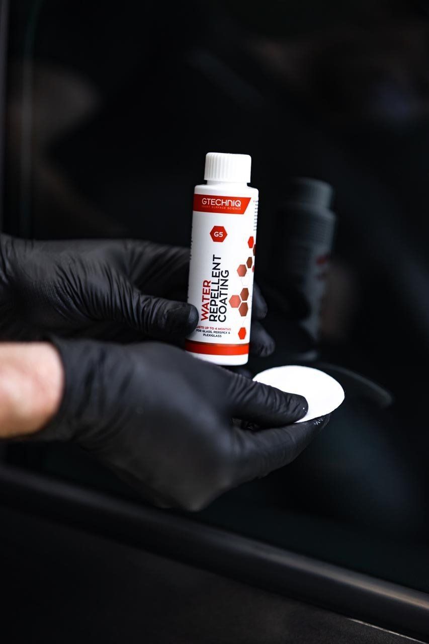 Person in black gloves holding a bottle of detailing product and applicator pad, against a black background.