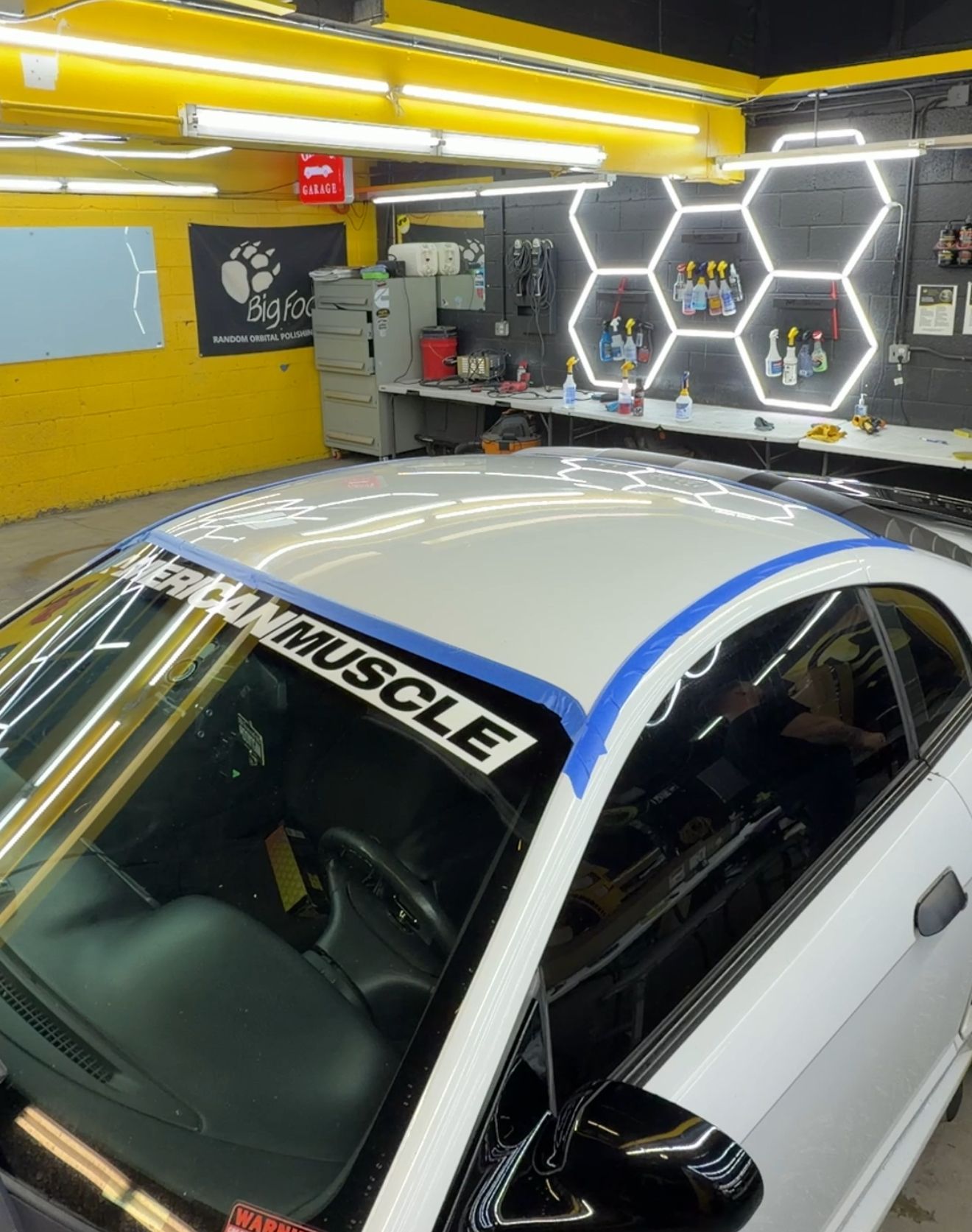 White car roof with blue tape, inside a well-lit workshop with hexagon lights and tools.