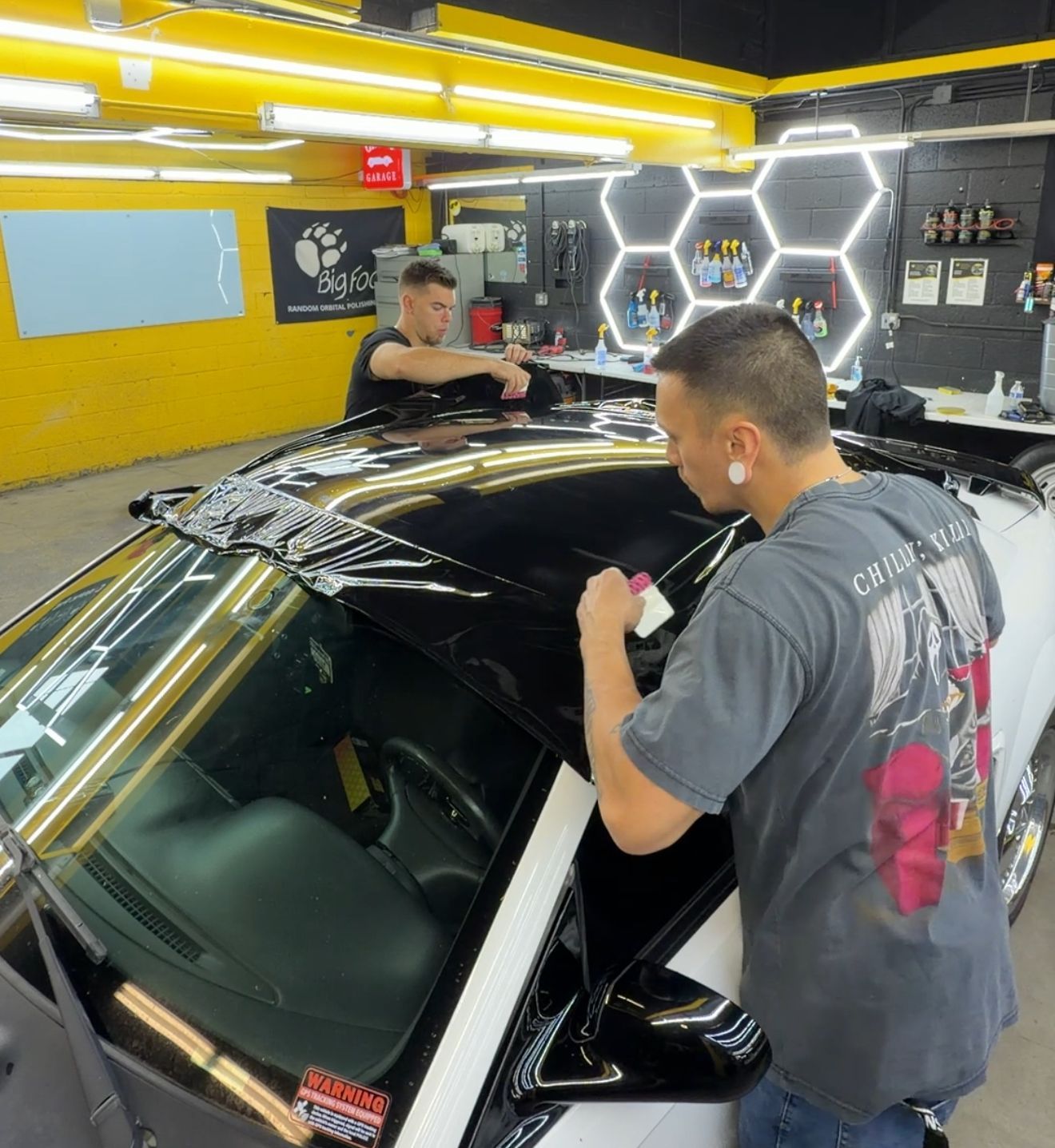 Two people applying black vinyl wrap to a white car roof inside a shop with yellow walls and hexagon lights.