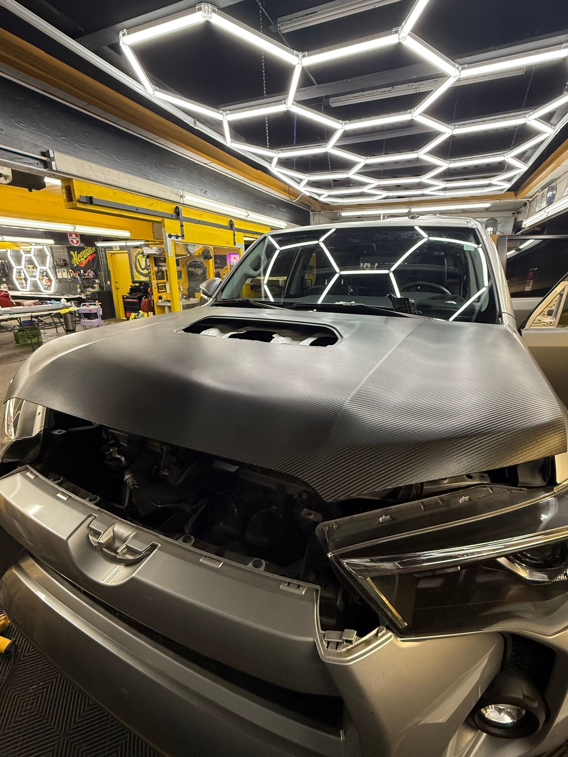 White car roof with blue tape, inside a well-lit workshop with hexagon lights and tools.