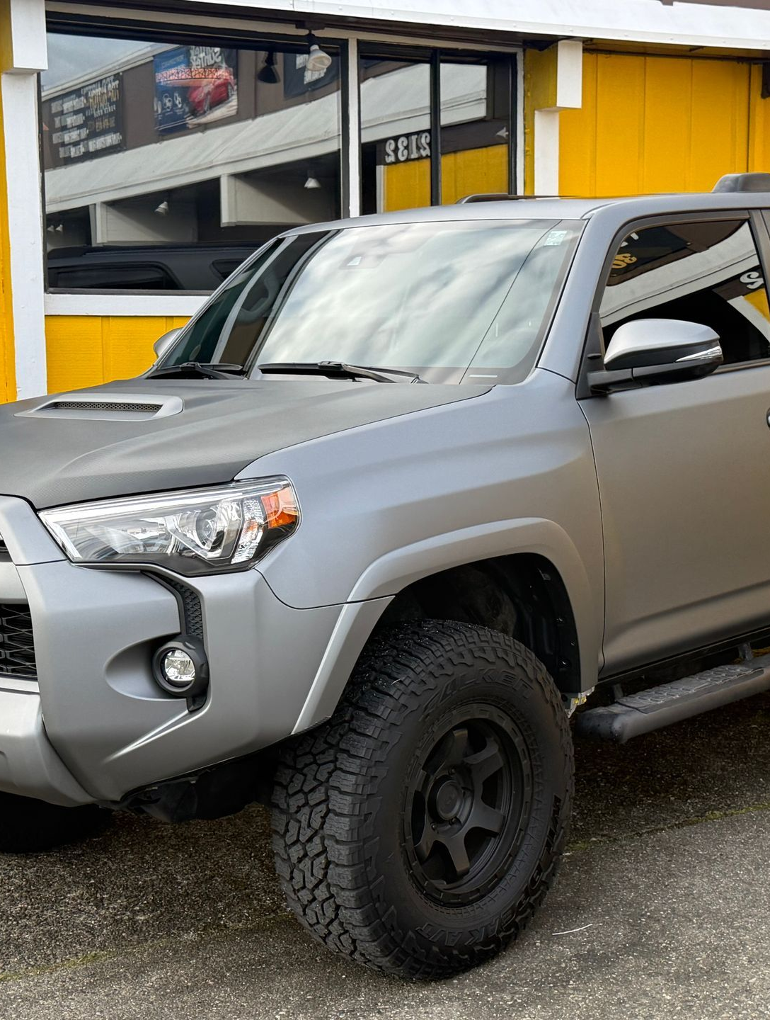 Two people applying black vinyl wrap to a white car roof inside a shop with yellow walls and hexagon lights.