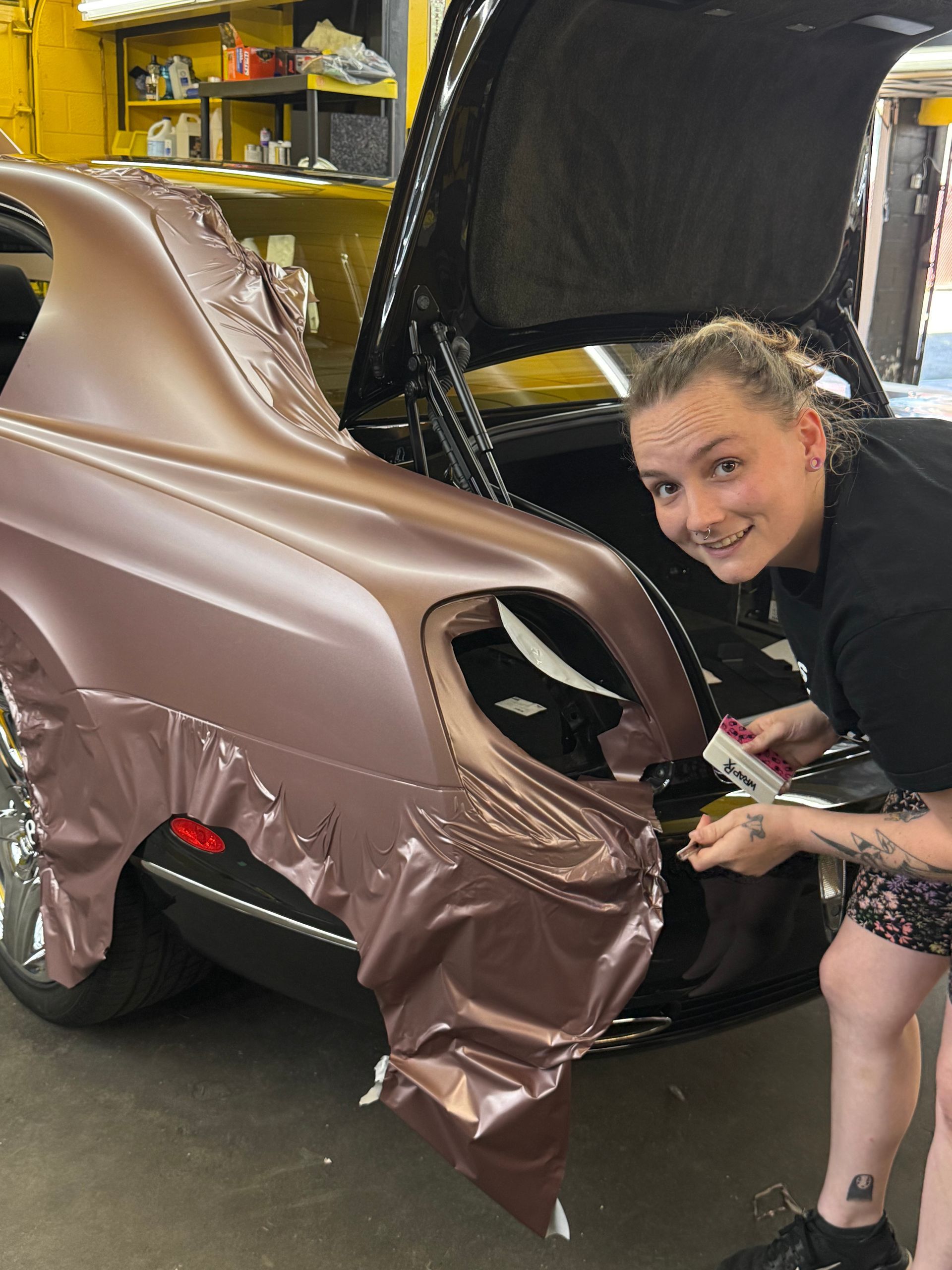 Woman wrapping a car's rear with brown vinyl in a workshop. Smiling, using a tool. Car is black with chrome wheels.