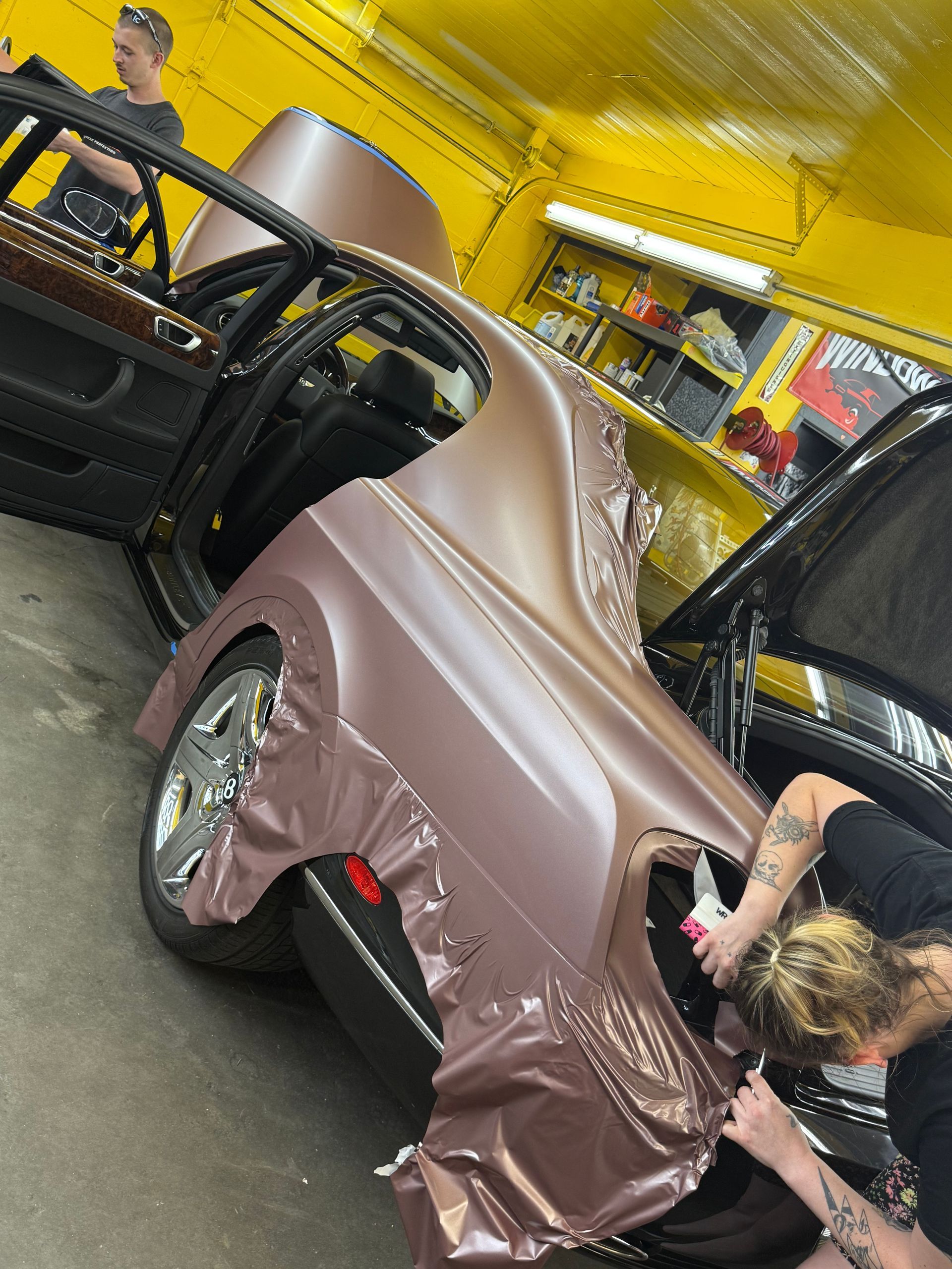 People wrapping a car in a dusty rose vinyl in a yellow-walled shop; one person works on the car's rear panel.