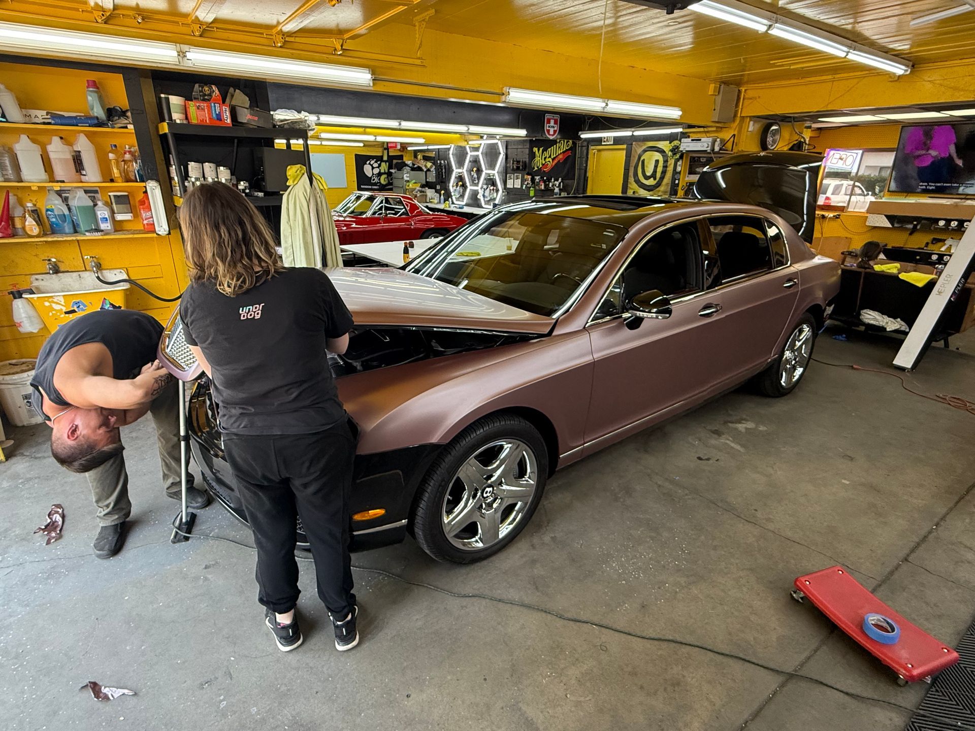 Two people installing vinyl wrap on a rose-colored Bentley in a garage.