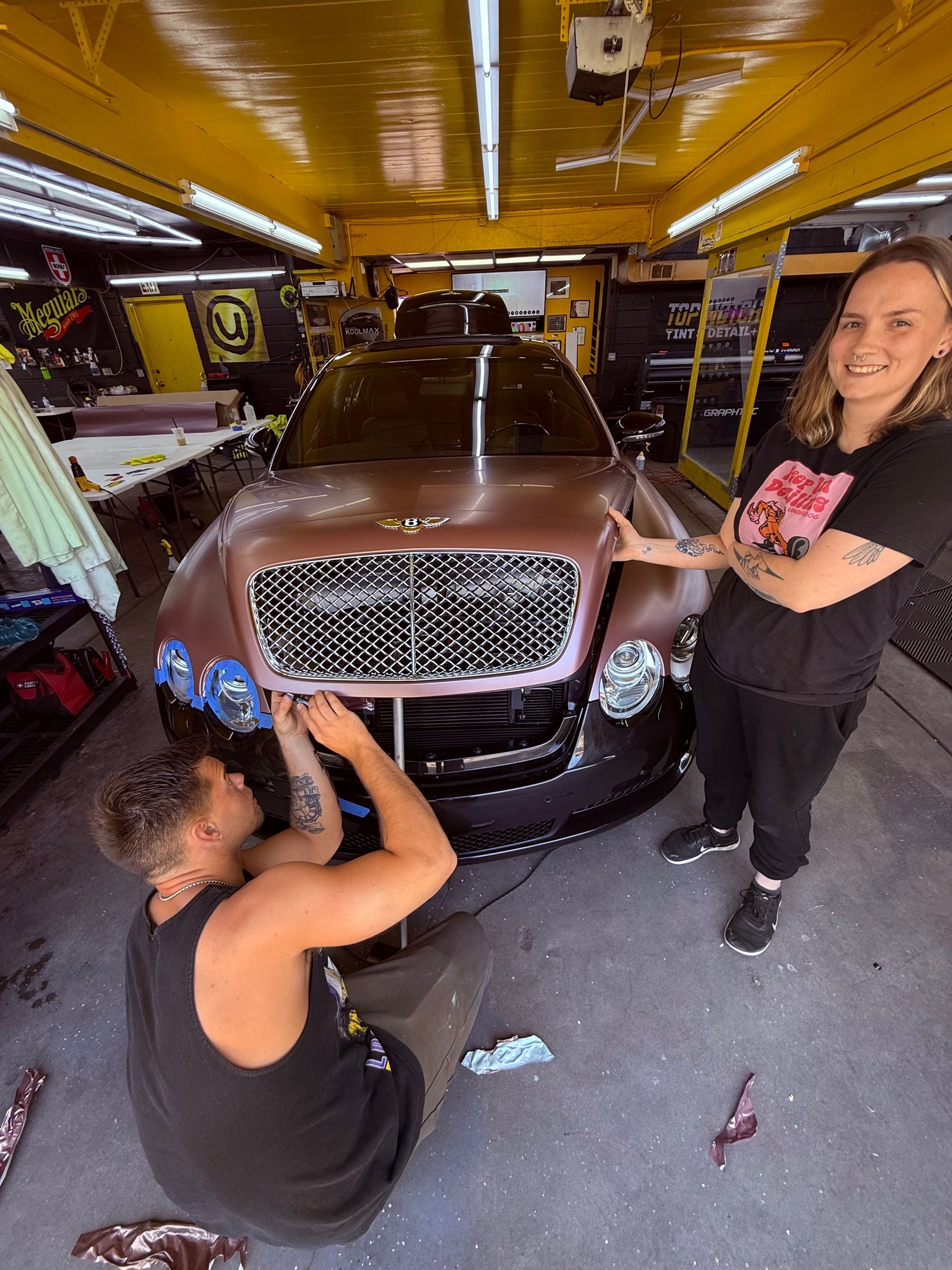 Two people wrapping a brown Bentley in a garage. One kneels, the other stands.