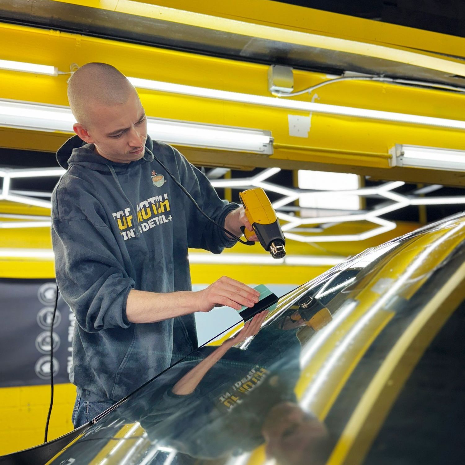 A man wearing a tie dye hoodie is working on a car windshield