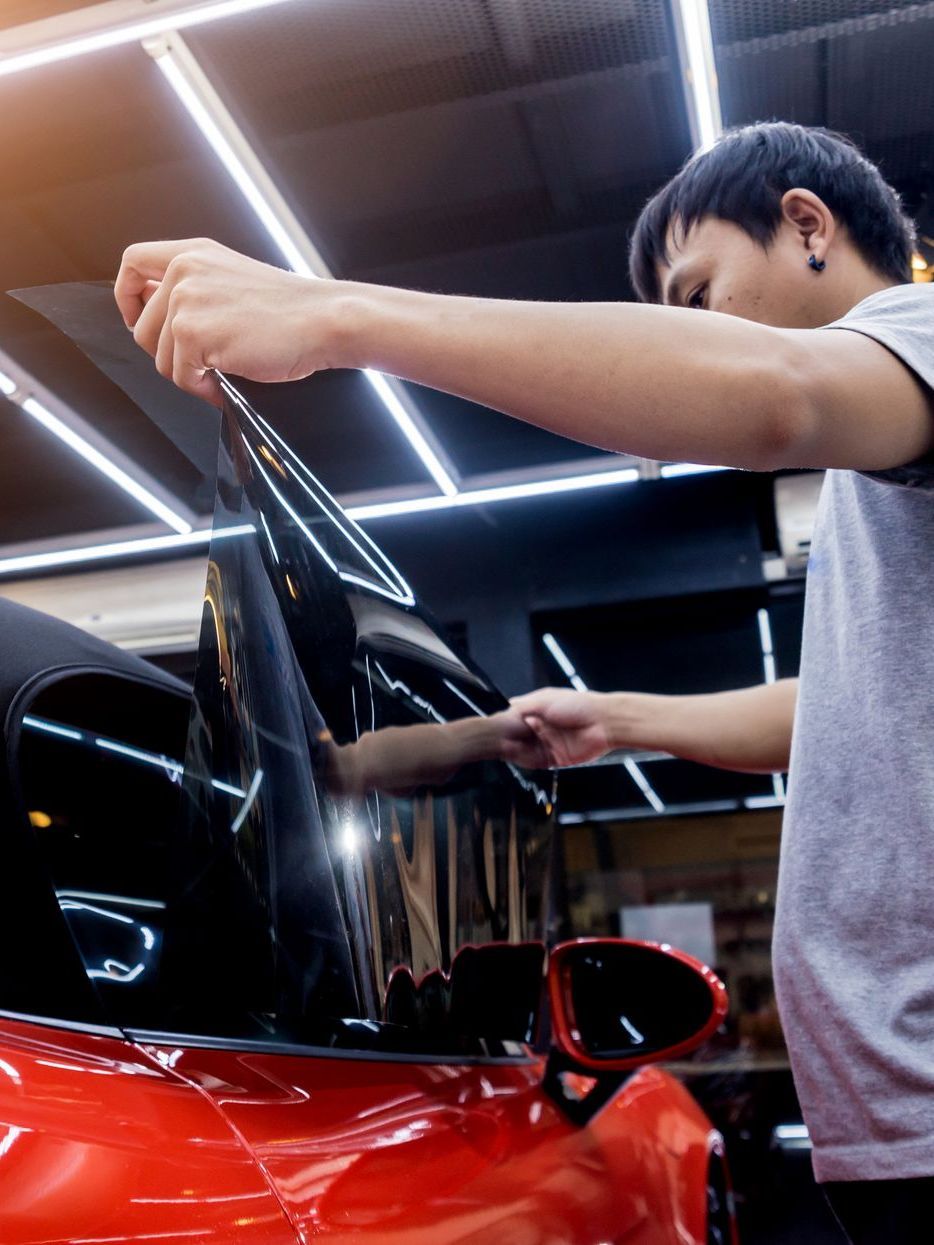 A man is applying window tinting to a red car in a garage.