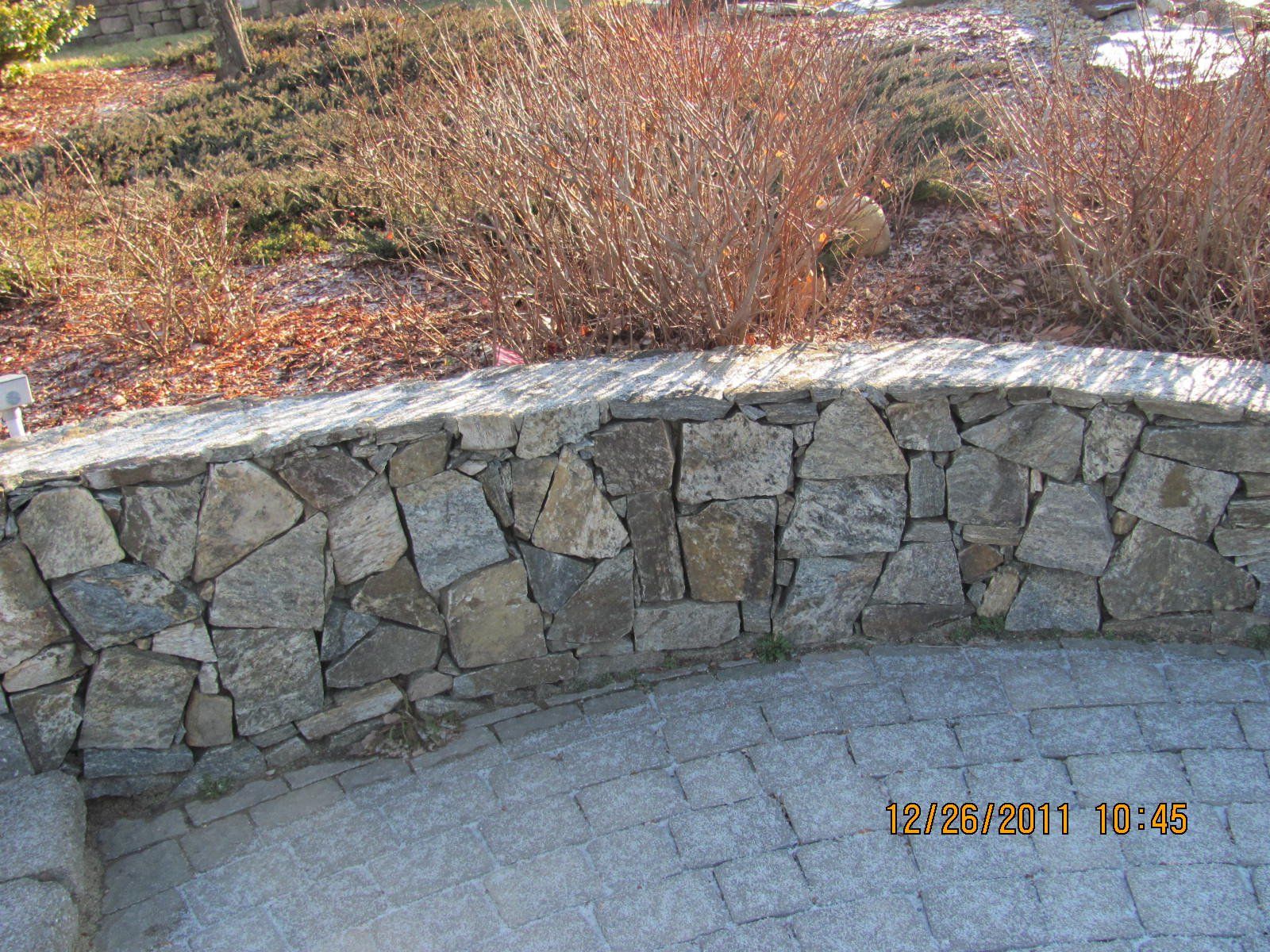 A stone wall is surrounded by bushes and a brick walkway.