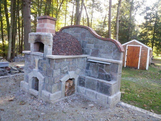 A stone oven is sitting in the middle of a yard next to a wooden shed.