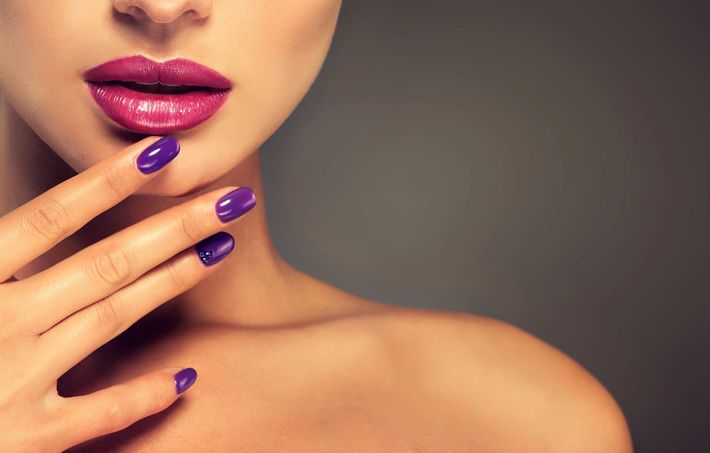 Primo piano di labbra rosa lucide e unghie curate viola su uno sfondo grigio tenue.