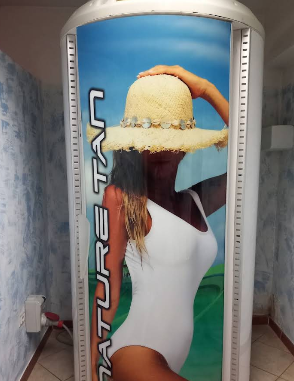Persona in costume da bagno bianco e cappello di paglia all'interno di una cabina abbronzante, con uno sfondo blu a tema spiaggia.
