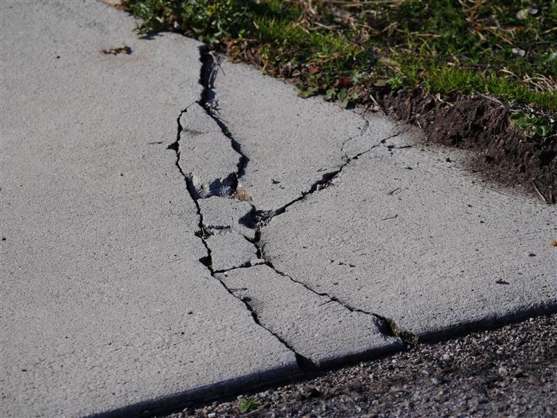 Cracked concrete sidewalk with large fissures, beside grass.