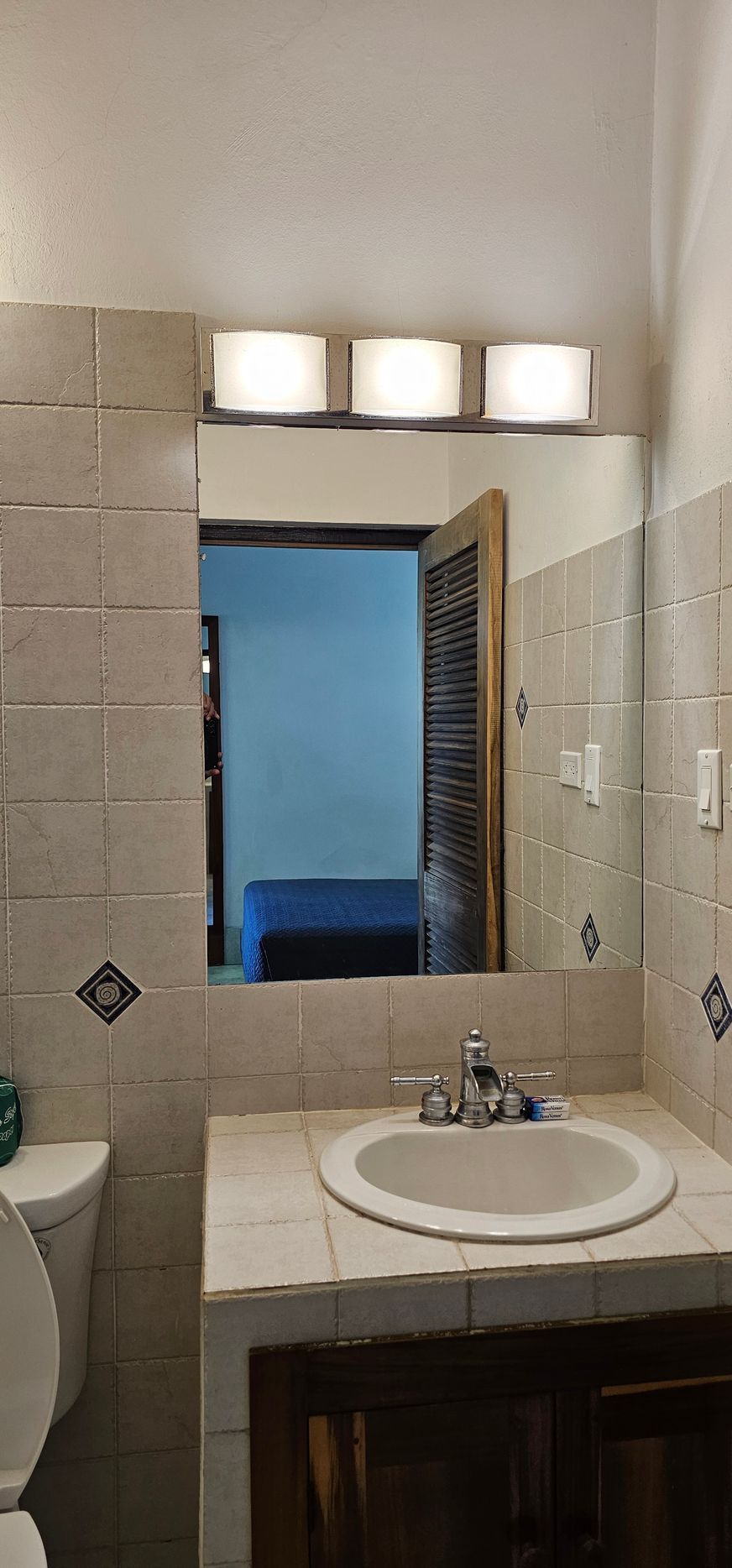 Bathroom with mirror, sink, and overhead lights. Tile walls and a door opening to another room are visible.