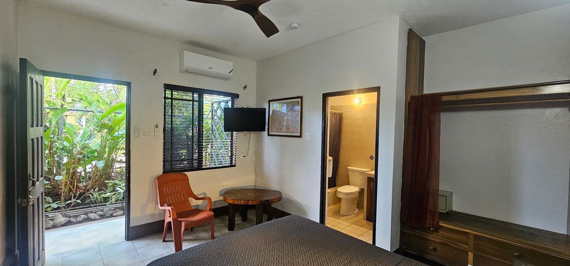 A bedroom with an open door to a garden, an orange chair, a table, a TV, and a door to a bathroom.