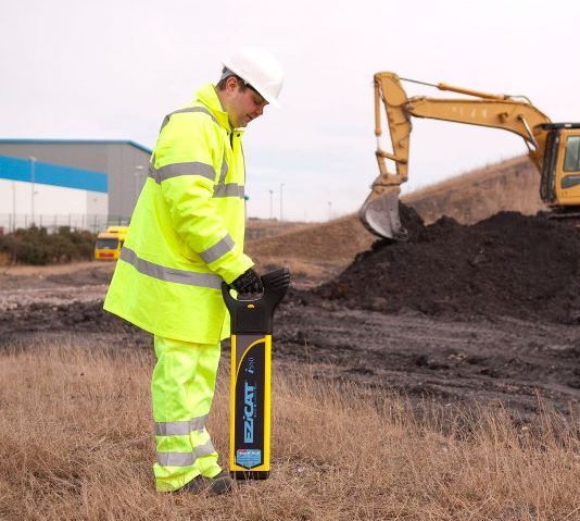 A man in a yellow jacket is standing in a field with a yellow excavator in the background