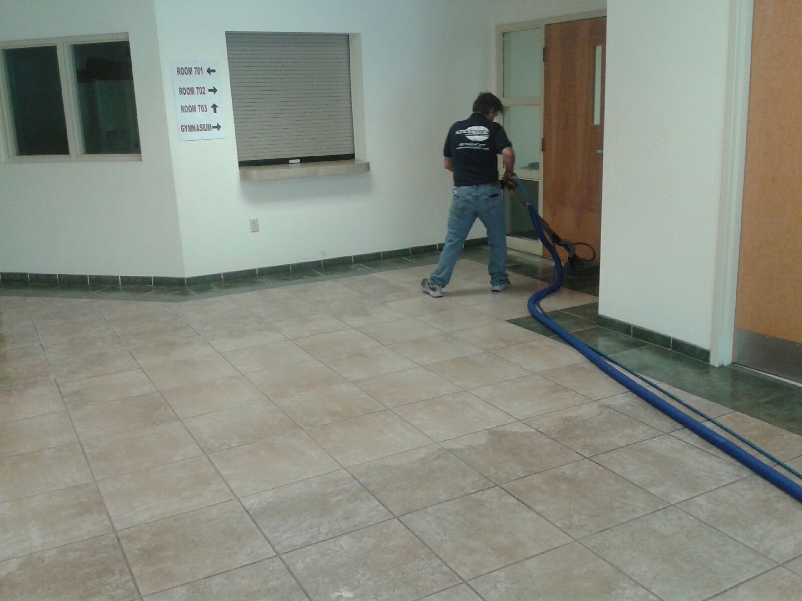 Worker vacuuming a tiled room with a blue hose near a window and doorway