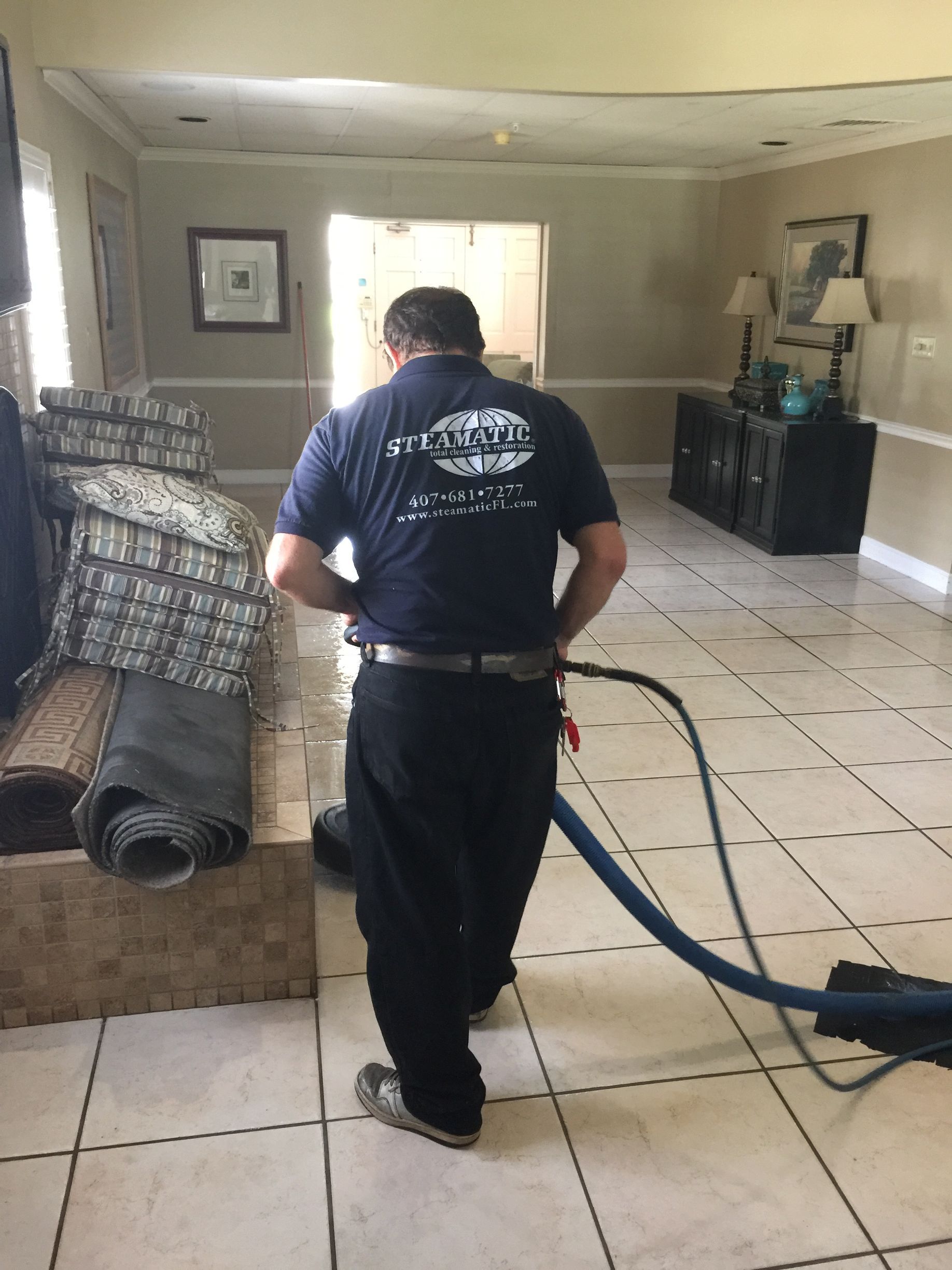 Worker using a large hose to clean a tiled living room with a couch and sideboard.