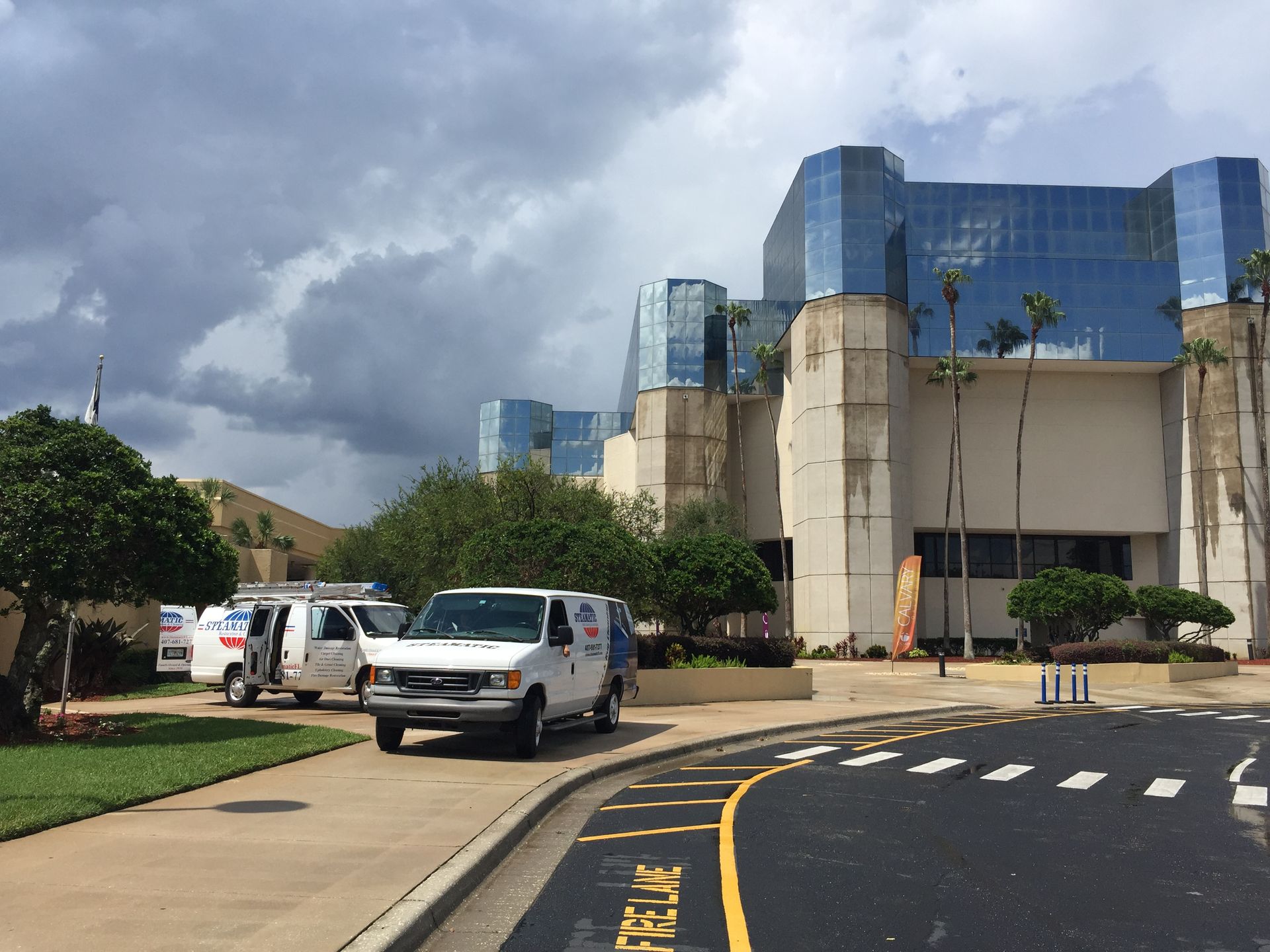 Modern building with blue glass facade, parked white vans, and cloudy sky by a curved driveway