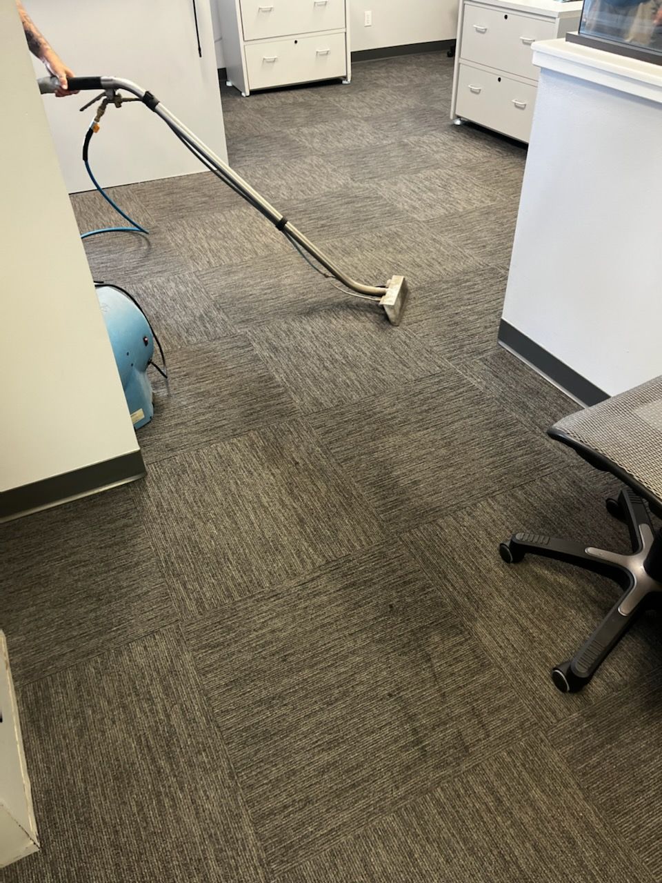 Person vacuuming a gray office carpet with a handheld vacuum near desks and a chair