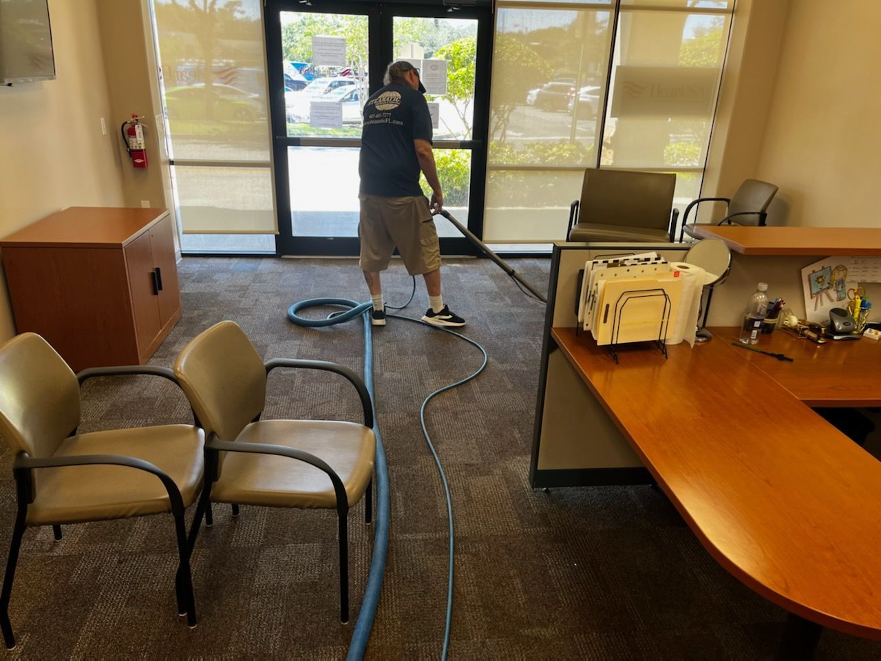 Person vacuuming an office lobby with chairs, desks, and glass doors leading outside.