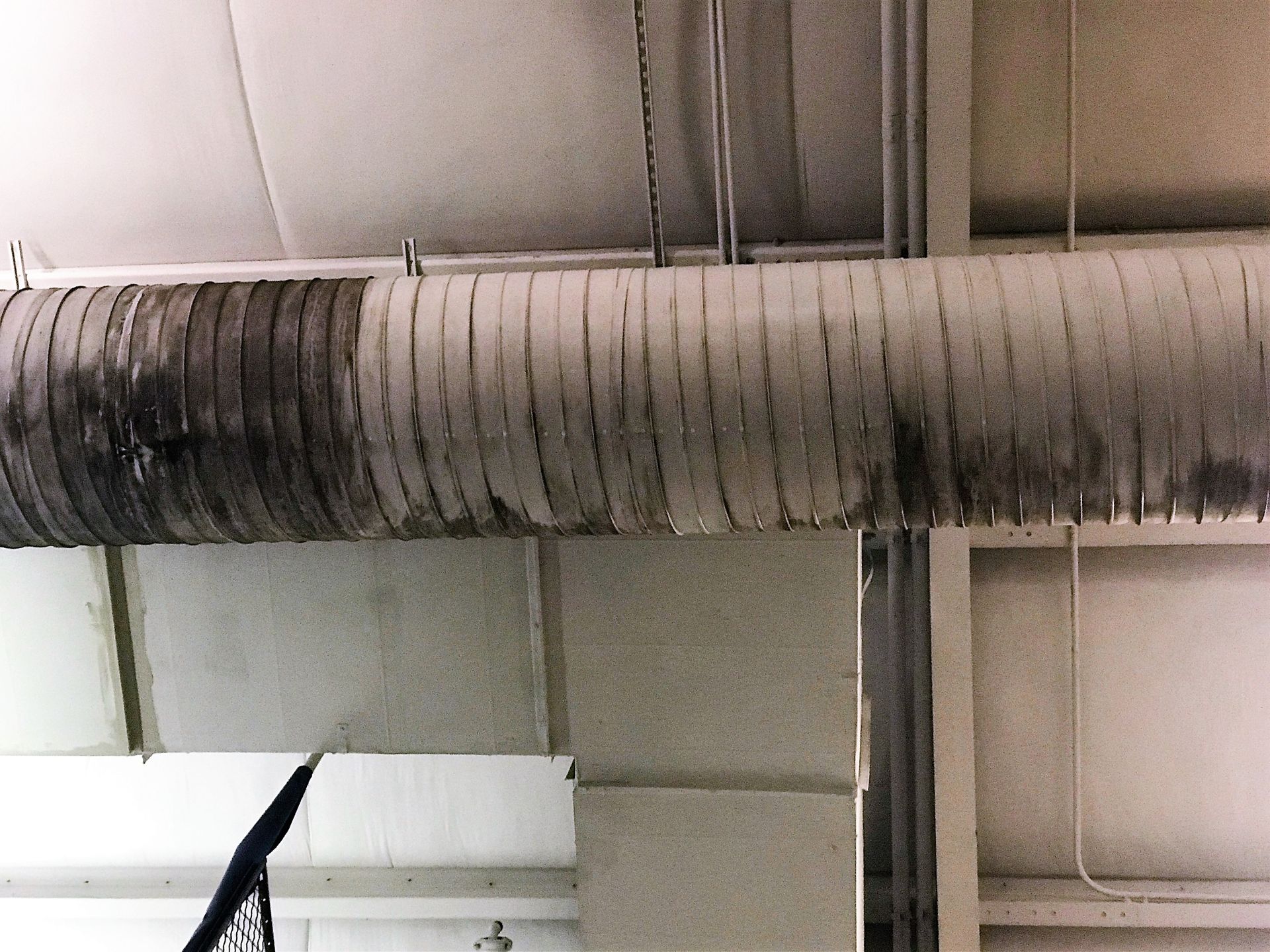 Dusty industrial ductwork on a ceiling with dark soot stains and pipes nearby