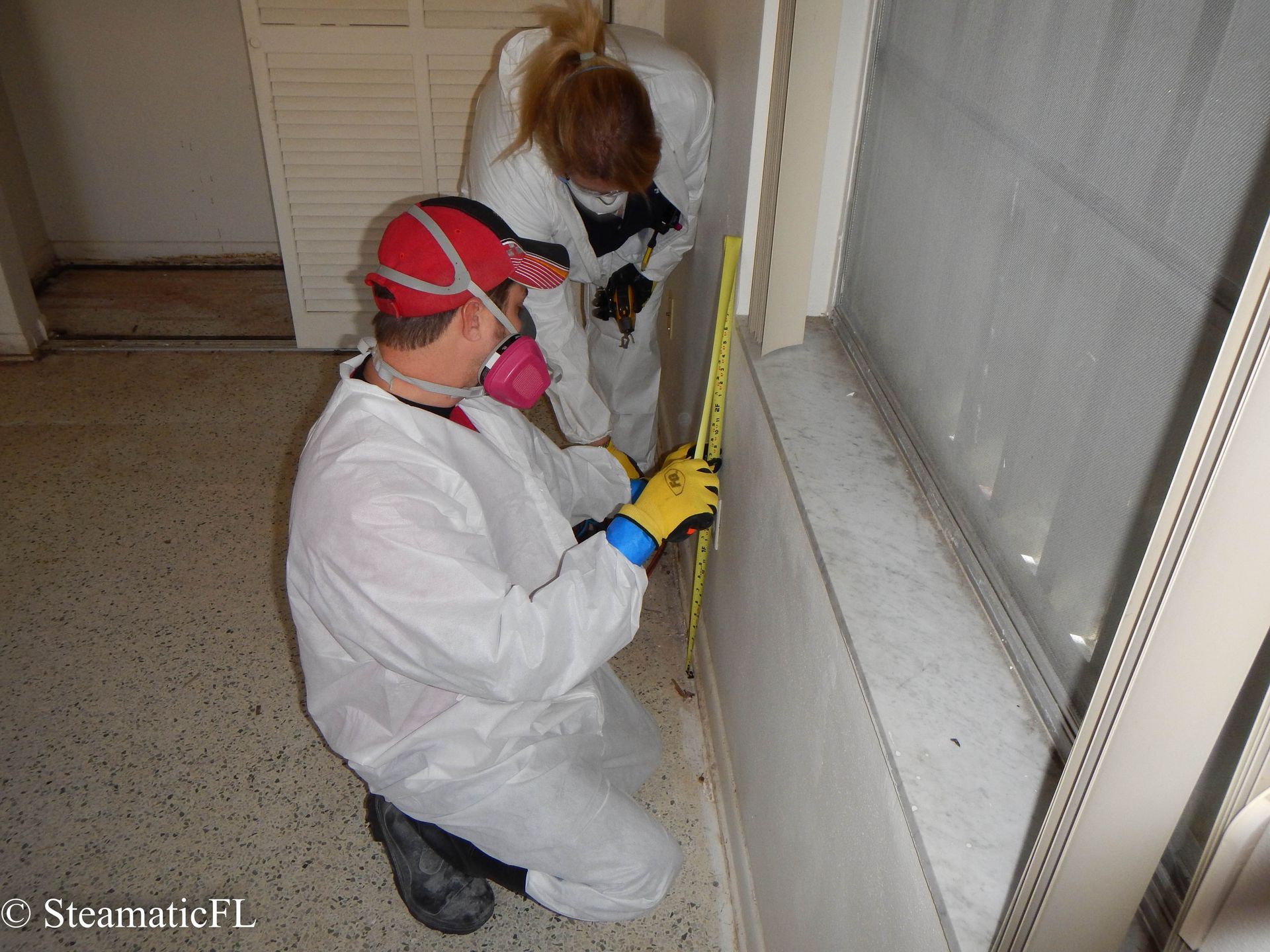 Two workers in white suits inspecting a wall with a tape measure in a narrow hallway