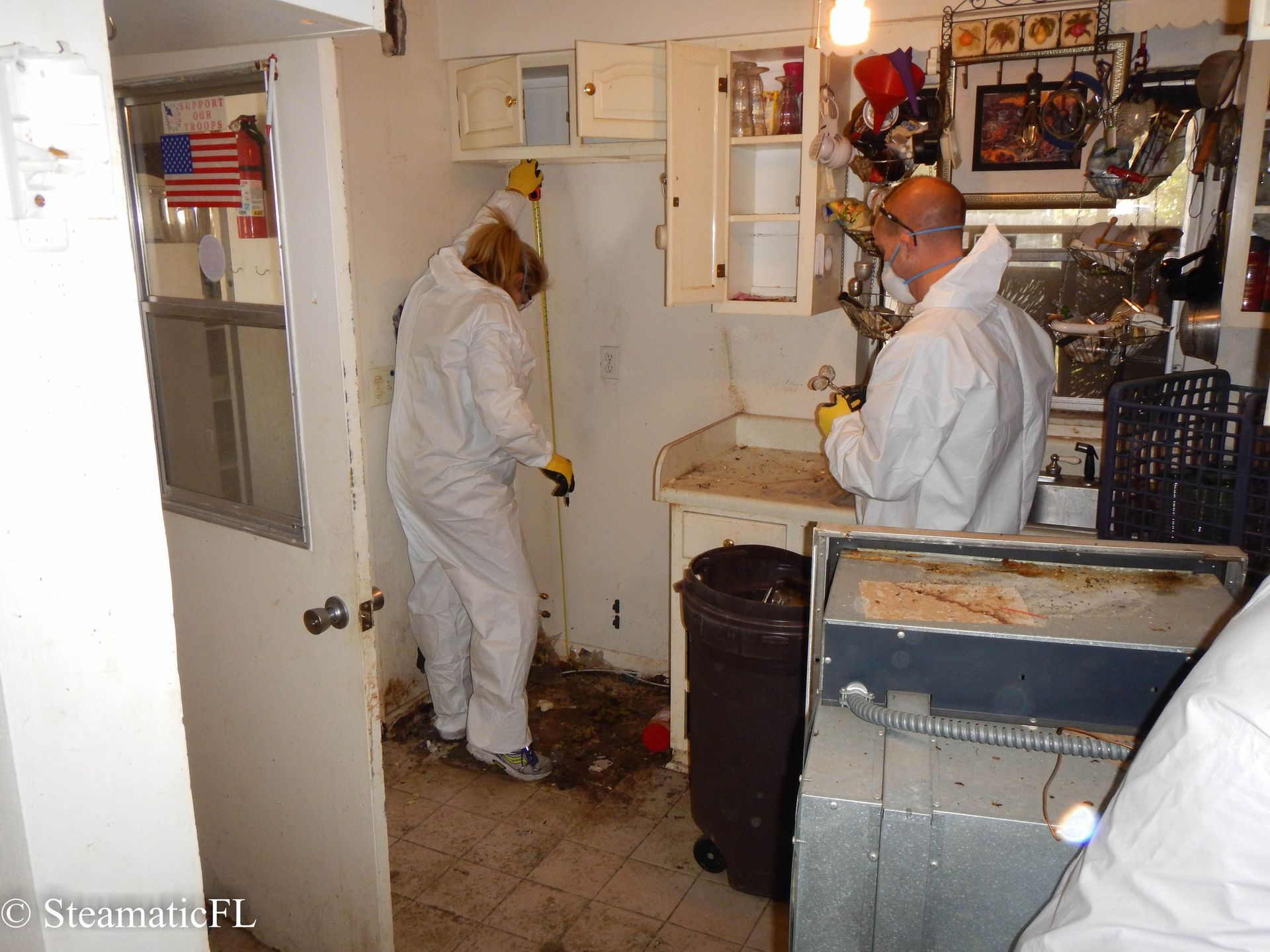 Two workers in white coveralls inspect a cluttered utility room with open cabinets and equipment.