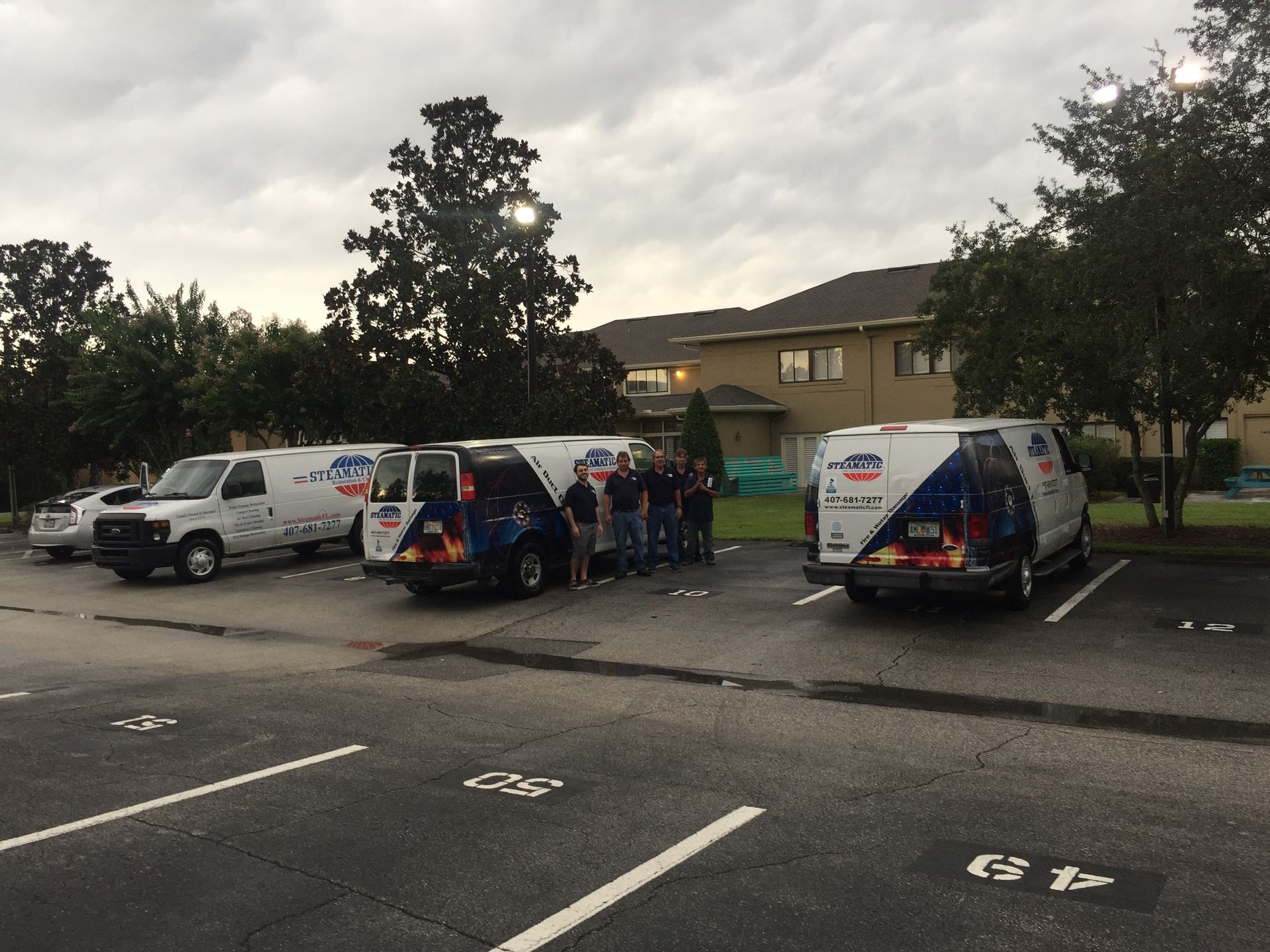 Parked vans and a small group of people in a lot outside a building at dusk.