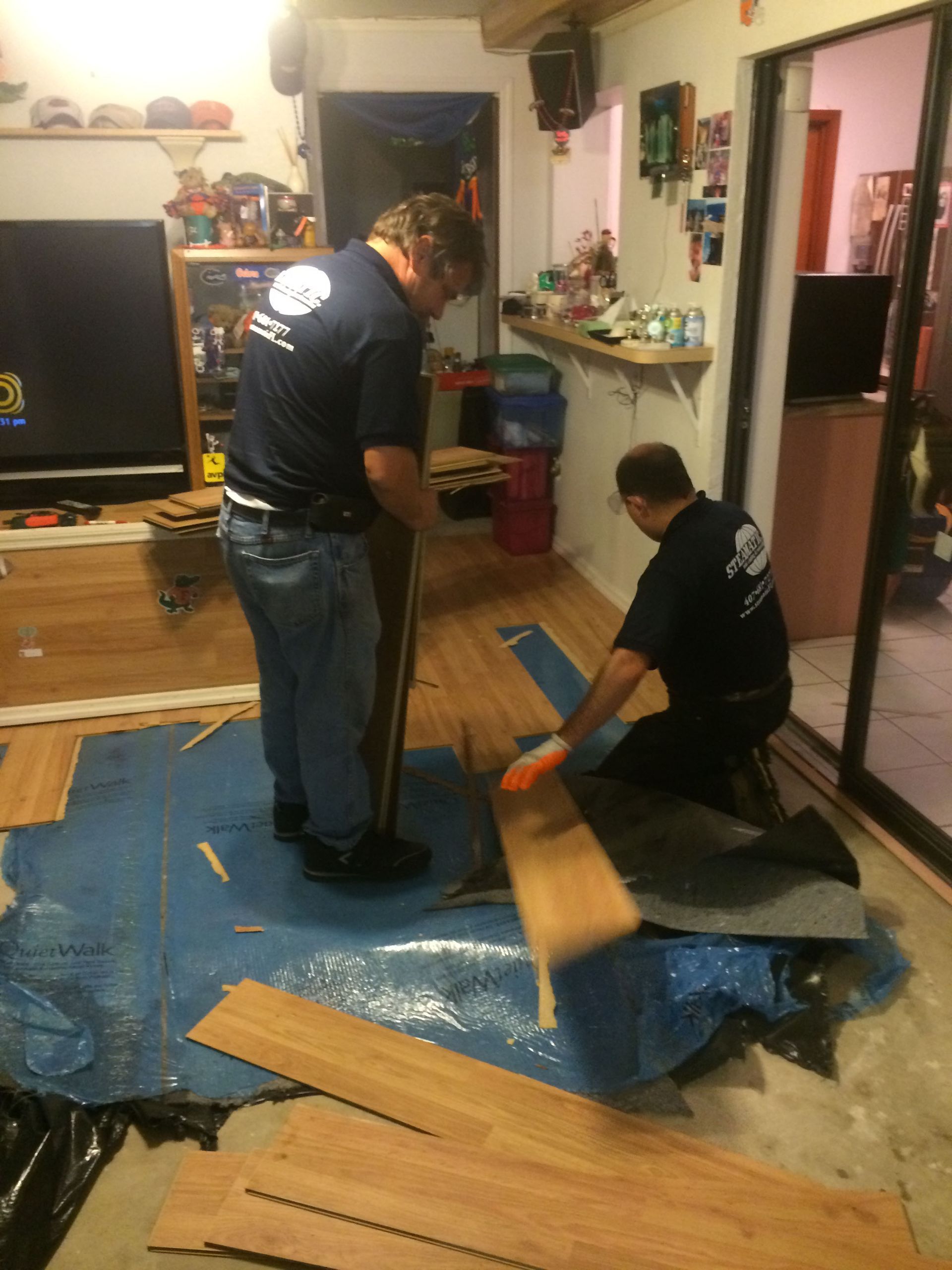 Two people installing wood flooring in a living room, one kneeling on blue tarp.