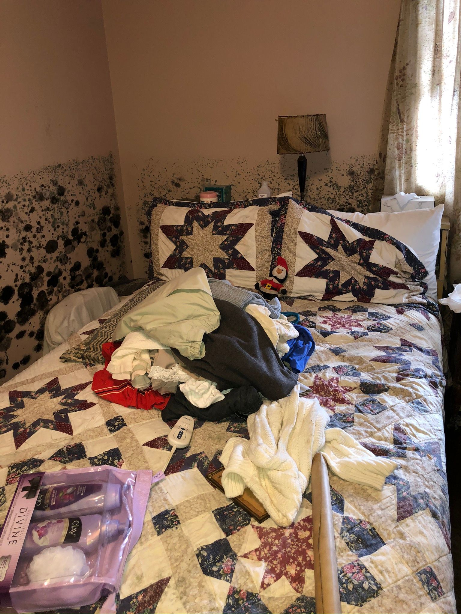 Bedroom with an unmade bed covered in clothes and patterned quilts, beside a bright window