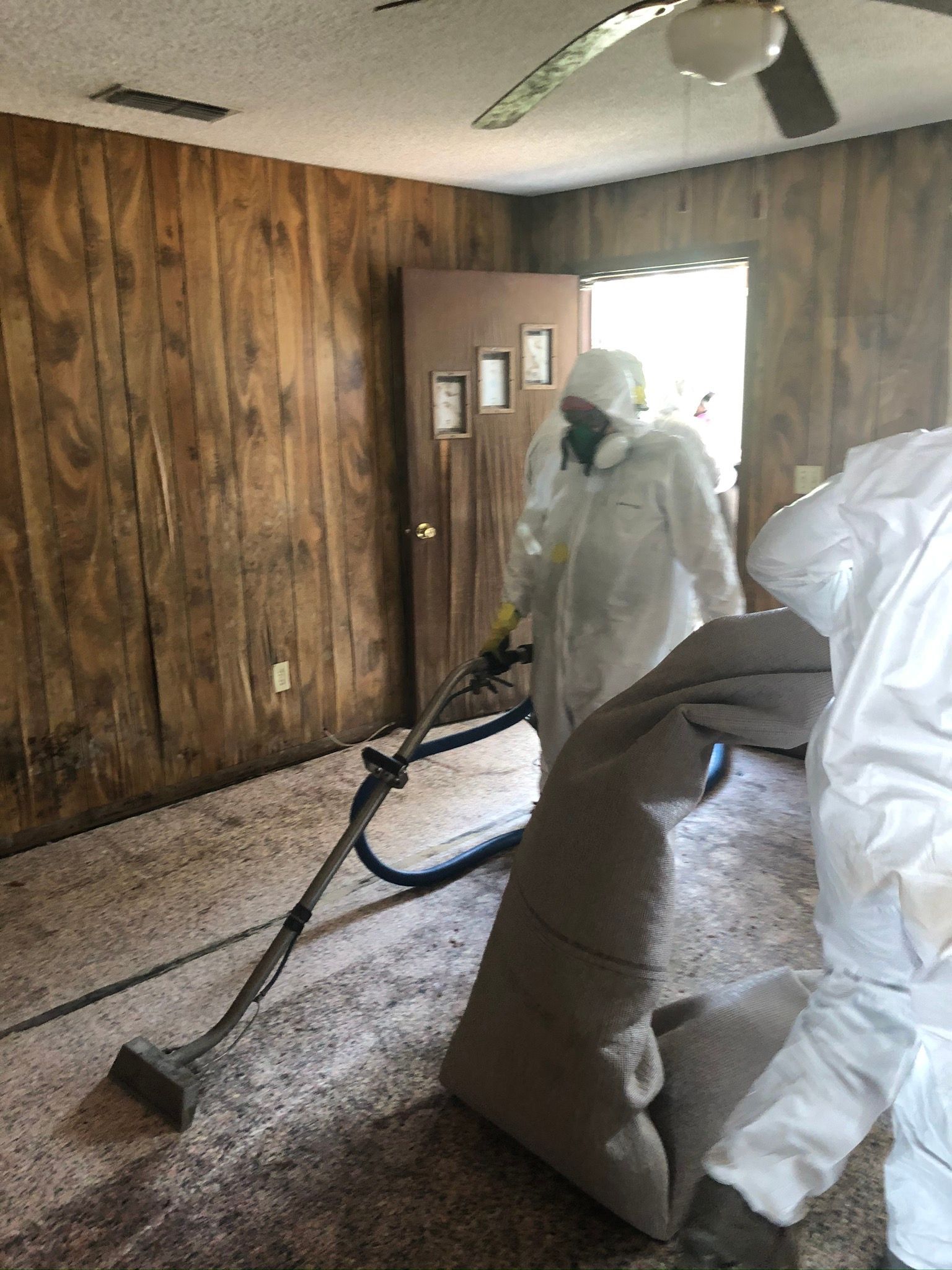Person in protective suit vacuuming debris in a damaged room with wood-paneled walls and piled plastic-wrapped items.