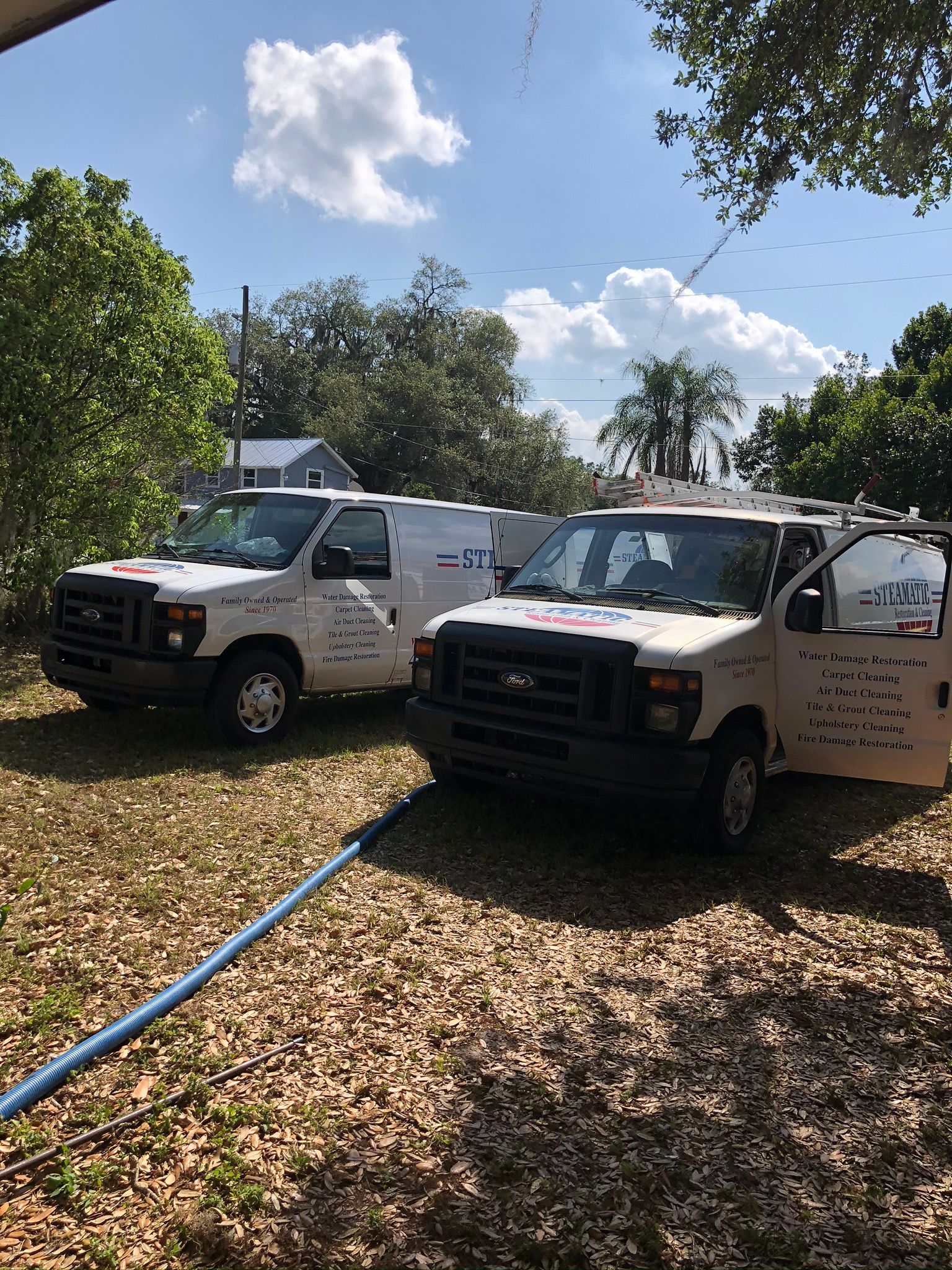 Two white utility trucks parked on leaf-covered ground beside a blue hose in a sunny wooded yard