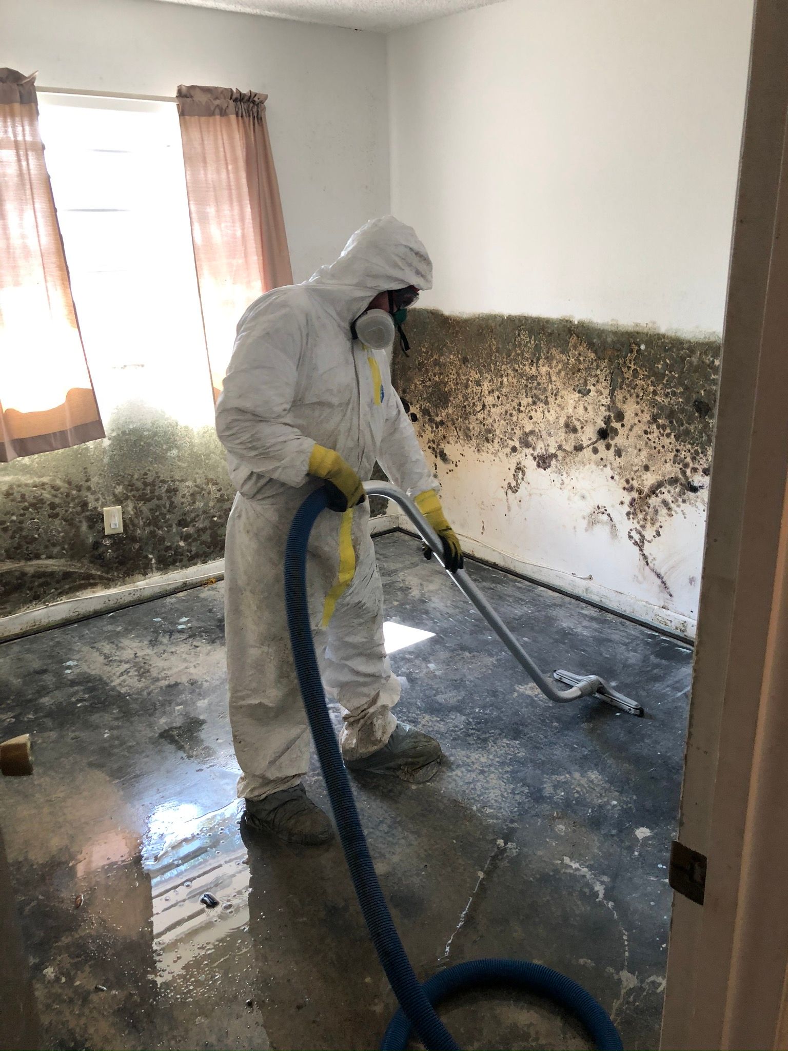 Worker in white protective suit cleaning a mold-damaged room with a wet vacuum and hose
