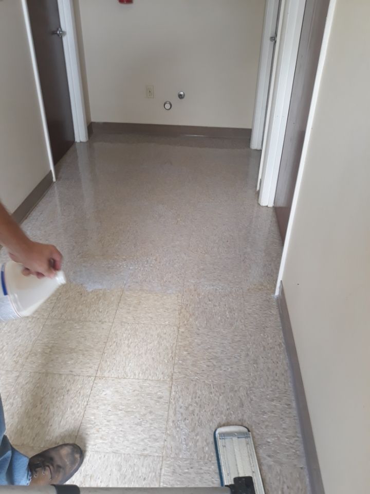 Empty hallway with tiled floor, open doorways, and a person’s hand holding a spray bottle at the left edge