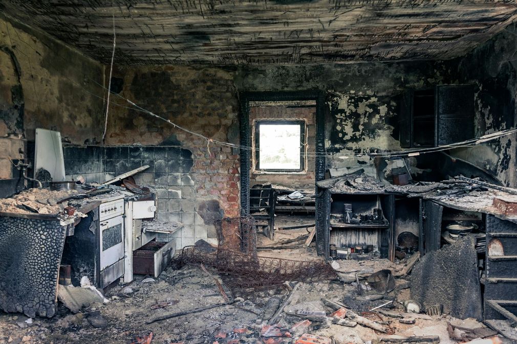Burned, gutted room with charred debris, damaged furniture, and a bright window in the center