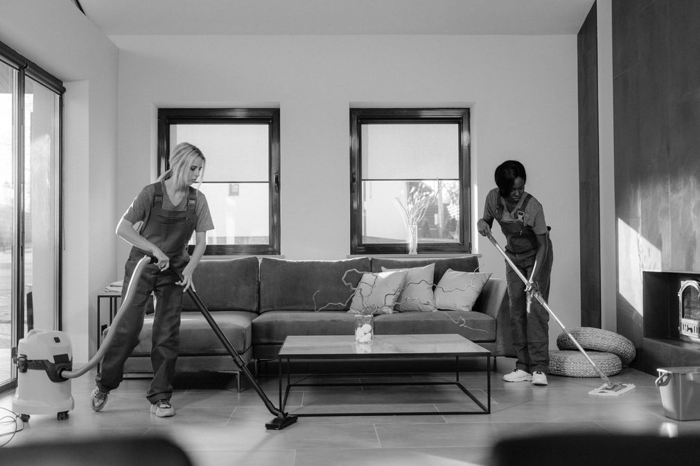 Two people vacuuming a bright living room with a sofa and large windows