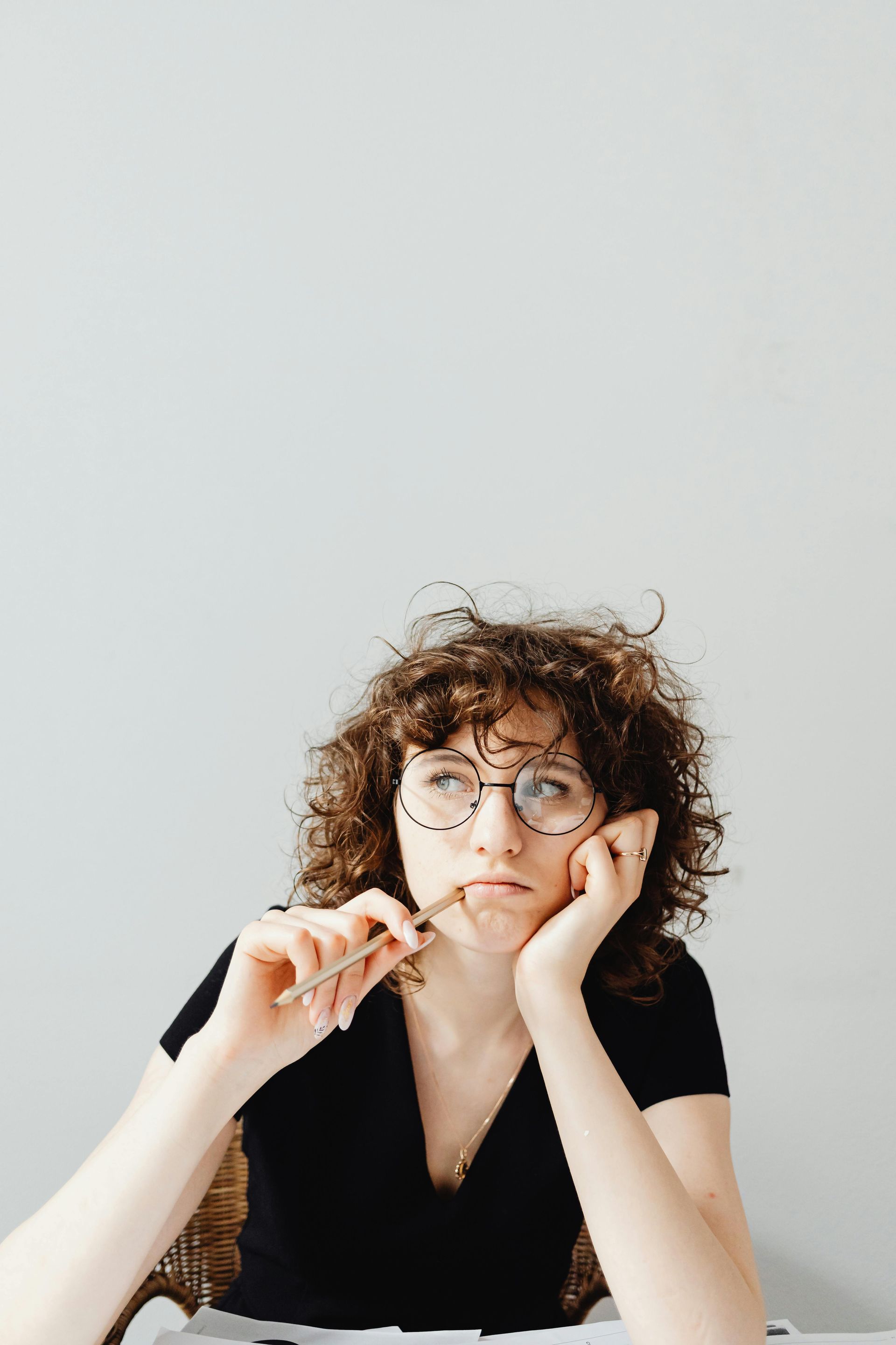 Person with curly hair and glasses resting their face on their hands, looking thoughtful against a plain wall.