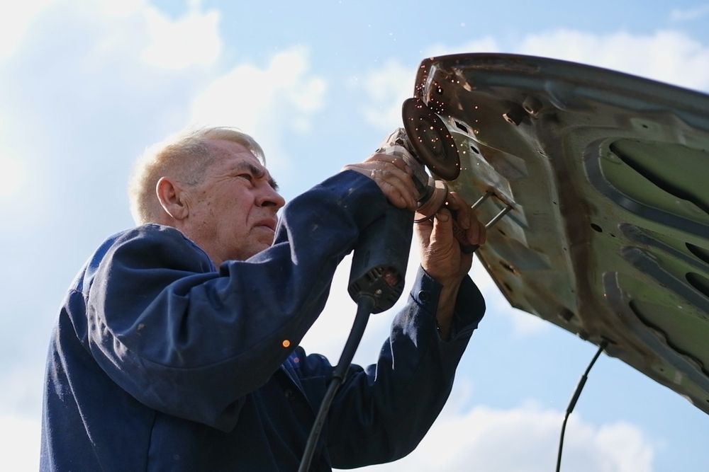 Man inspecting the underside of a car hood outdoors against a blue sky