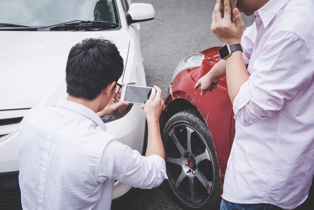 Two men at a fender bender, one taking a photo of damage to a red car, the other pointing at it.