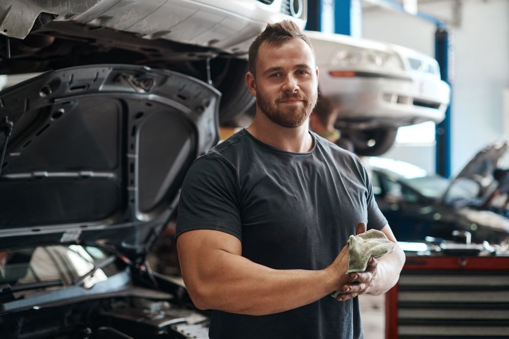 A man is standing in front of a car with the hood open in a garage.