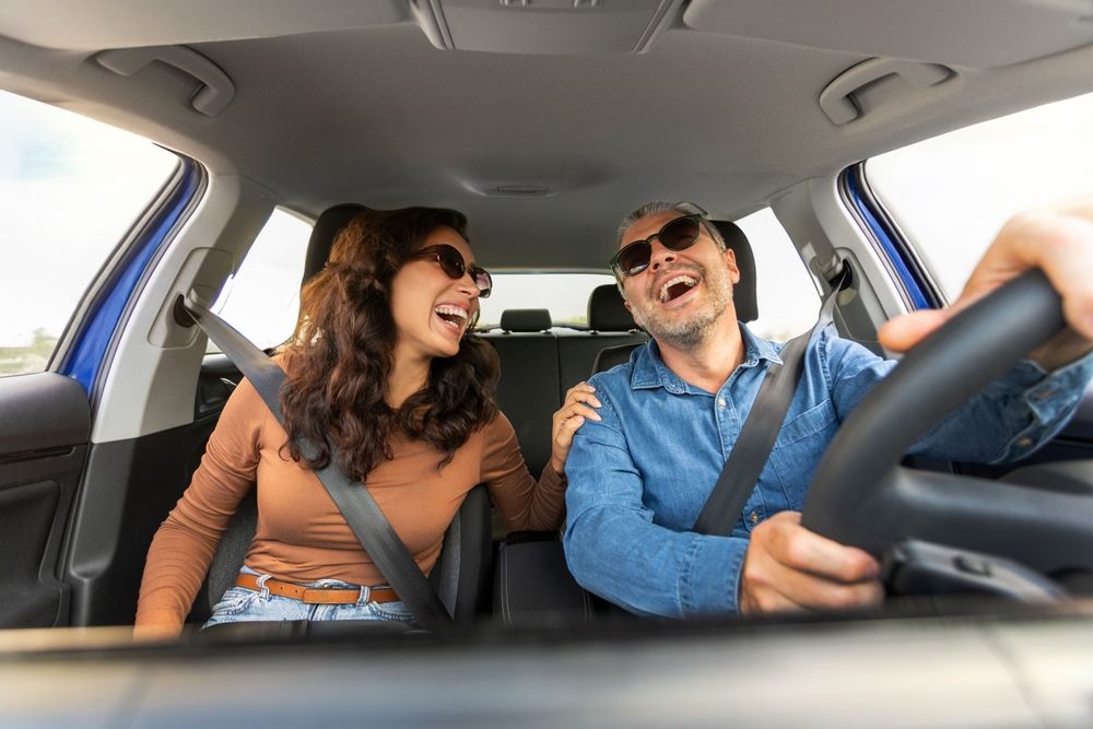 Couple in a car laughing, wearing sunglasses and seatbelts, with blue car interior and sky visible.