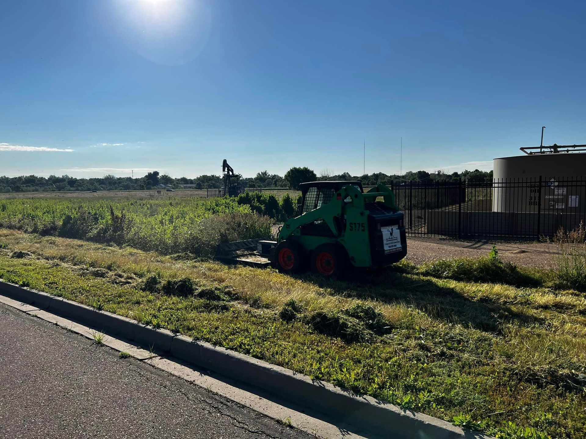 Green skid steer mower cutting grass next to a road, field, and clear sky.