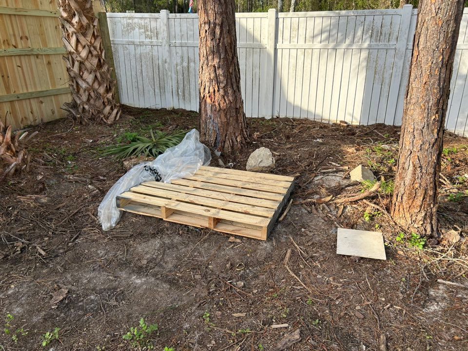 Wooden pallet and debris in a yard with trees and a white fence.
