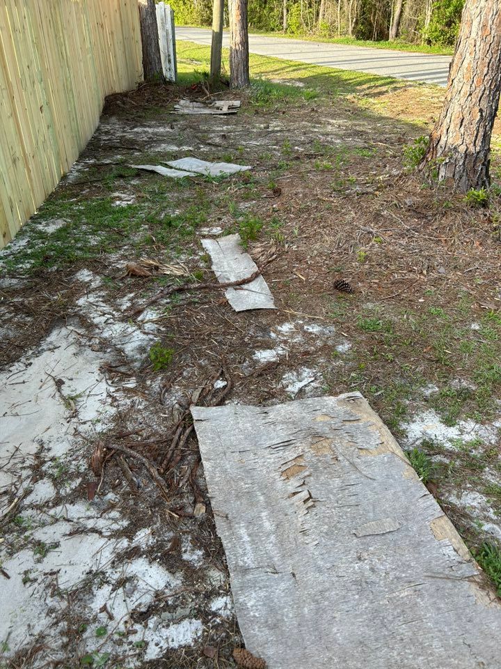 Dirt and sand path alongside a wooden fence. Gray planks lay on the ground, with sparse grass and trees nearby.