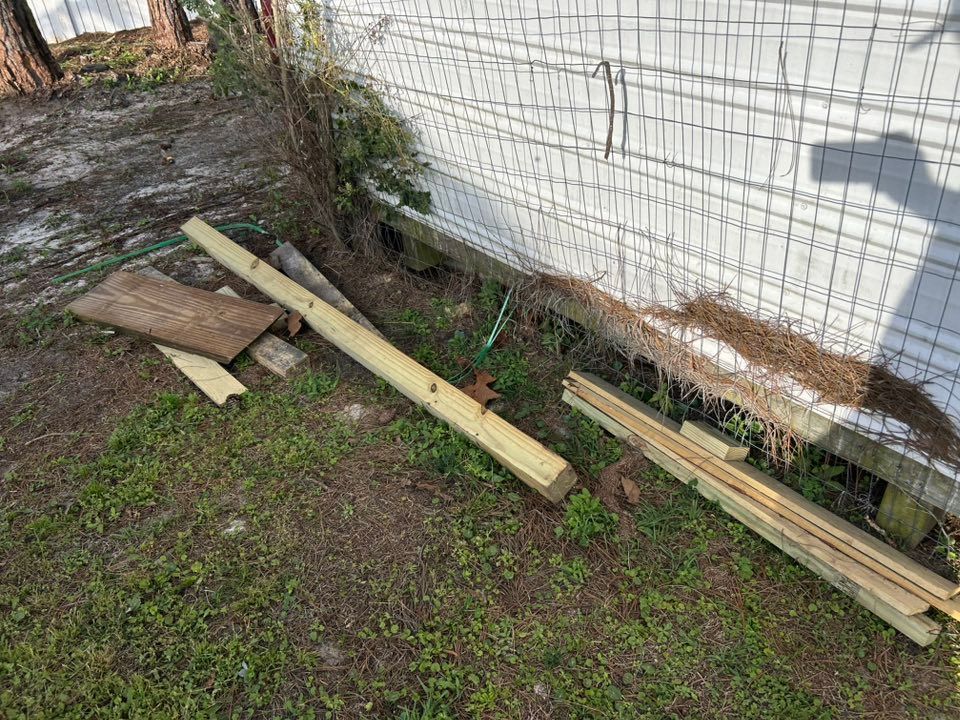 Wooden planks and debris scattered on grass next to a white wall with wire mesh.