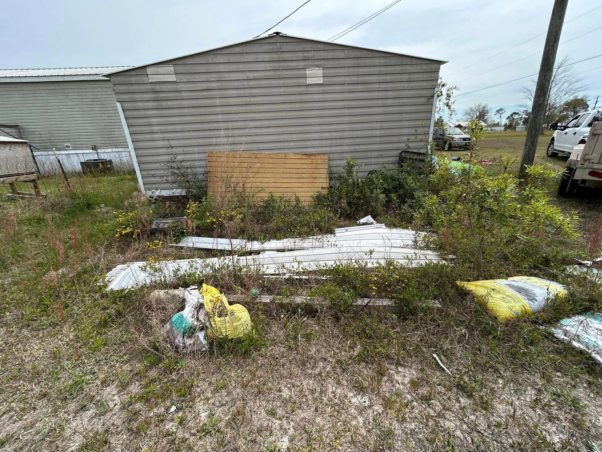 A weathered, gray building with debris in front, including boards and bags. Overgrown grass.