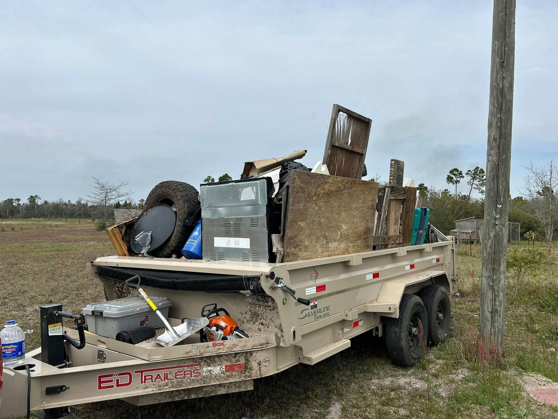 A loaded trailer filled with trash and debris parked next to a wooden utility pole in a field.