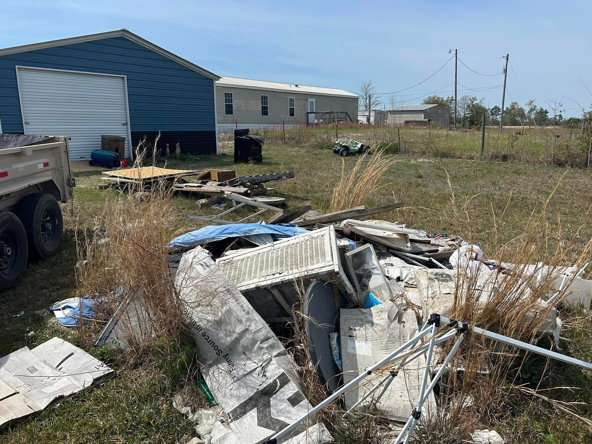 Pile of debris and trailer in a field with a house and mobile home in the background on a sunny day.