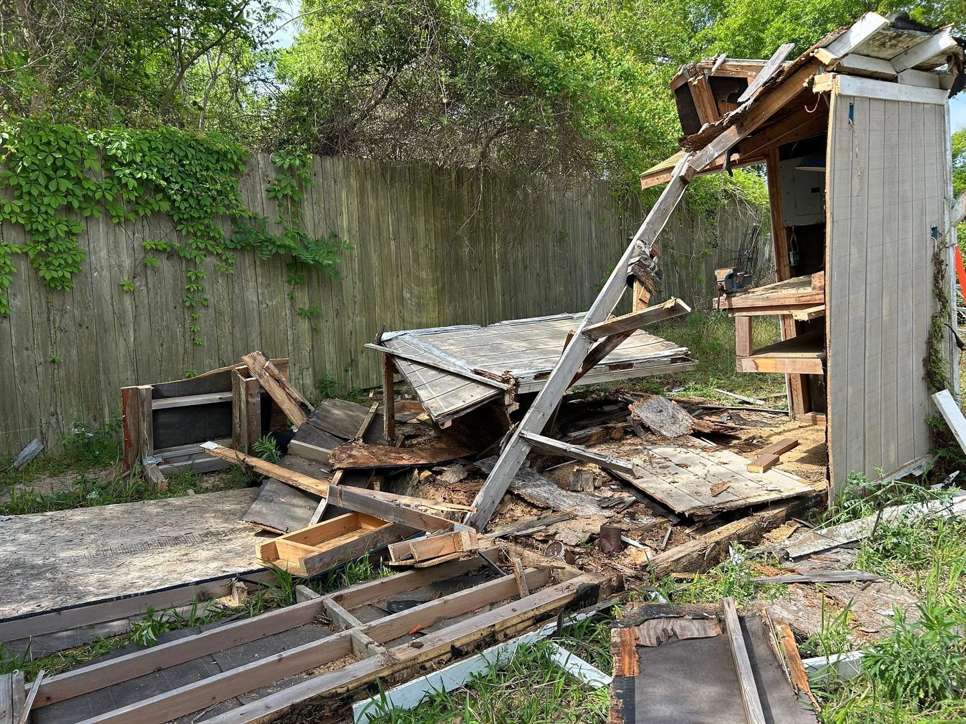 Collapsed wooden shed and debris in a yard, against a wooden fence with green foliage.